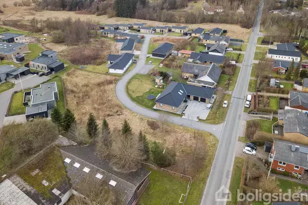 Boligområde med moderne huse ligger langs snoede veje, omgivet af grønne plæner og spredte træer. En hovedvej med parkerede biler ses til højre i billedet.