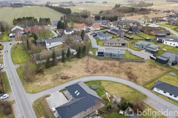 Luftfoto af en lille by med huse omgivet af marker og træer. Veje snor sig gennem kvarteret, der består af forskellige størrelser boliger. Området er grønt og landligt.