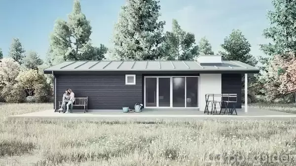 Et lille, mørkt træhus står på en åben græsplæne. En person sidder på en bænk ved siden af huset. Et bord og to stole er placeret på terrassen. Skovklædte træer ses i baggrunden.