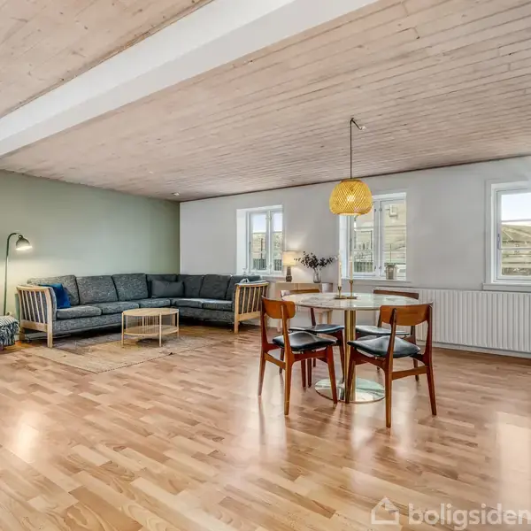 Stue med hjørnesofa til venstre, oplyst af en gulvlampe, står på trægulv ved grøn væg. Rundt spisebord med fire stole til højre, lyse vægge, loft og vinduer.