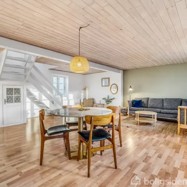 Stue med en rund spisebord og stole, indrettet på et trægulv. Sofa med sofabord står ved en grøn væg. Trappe fører opad. Loftslampe hænger over bordet. Lysindfald fra vinduer. Ingen tekst.