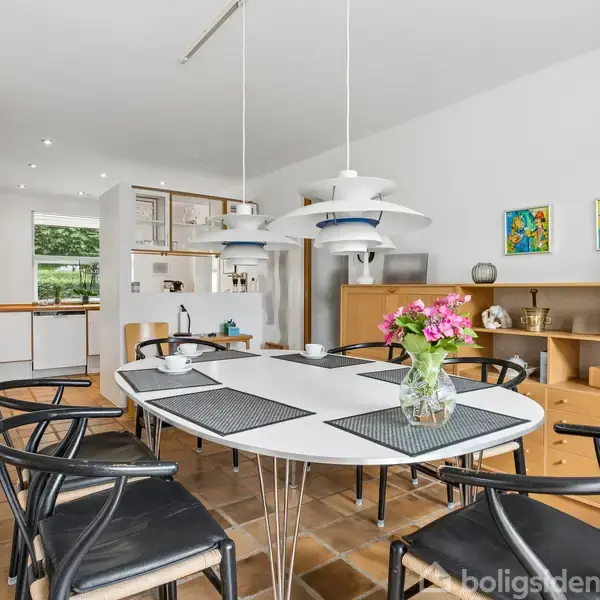 Ovalt spisebord med sorte stole står i moderne spisestue. Der hænger tre hvide lamper over bordet. I baggrunden er der hylder med dekorationer og farverige malerier.