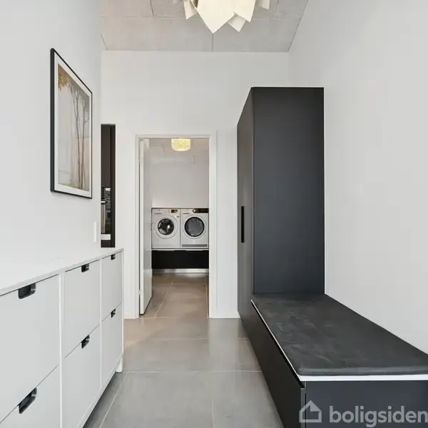 En moderne gang med hvidt kabinet og sort bænk fører til et vaskerum med vaskemaskine og tørretumbler. Billeder hænger på væggen, og rummet er lyst og rummeligt.