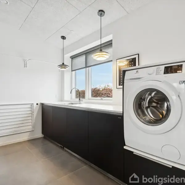 Vaskemaskine og tørretumbler er monteret på væggen. Rummet er lyst, moderne og indeholder mørke underskabe ved et vindue. Tøjstativer og en radiator ses også.