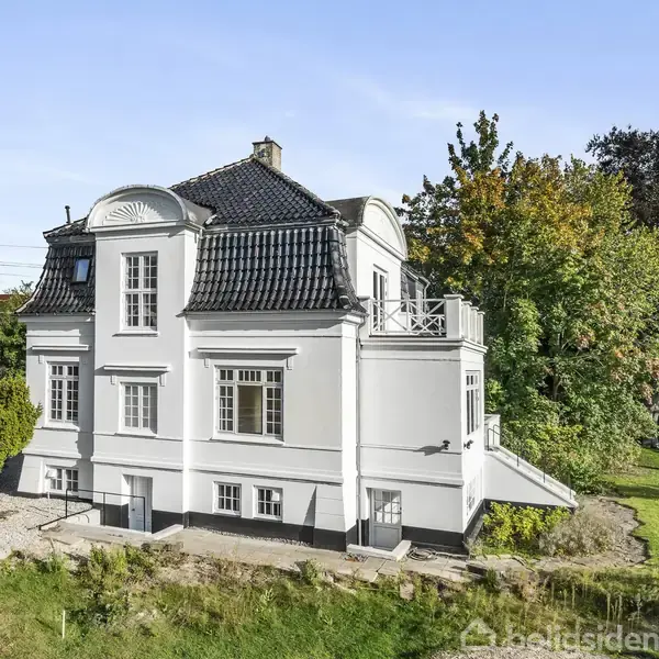 En hvid bygning med sort tag står i en grøn have med træer. Bygningen har flere vinduer og en udendørs trappe. Det er solskin, og himlen er blå.