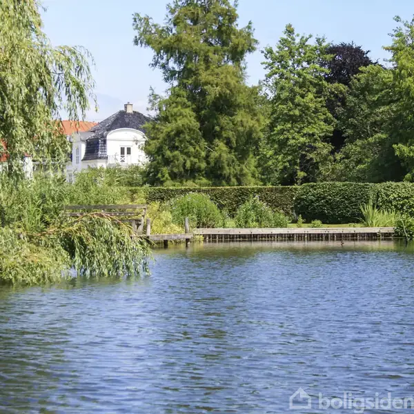 En sø med klart vand omgivet af tæt bevoksning og træer; en båd ligger ved en lille bro, mens et hvidt hus med rødt tag er synligt i baggrunden.