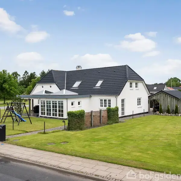 Hvidt to-etagers hus med sort tag ses med velplejet græsplæne og gyngestativ. Skilt med "Skole" står tæt ved, omgivet af grønne træer og blå himmel i et roligt kvarter.