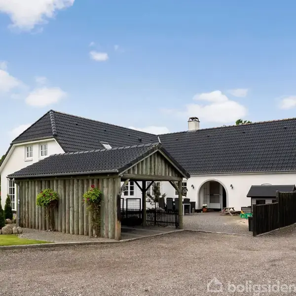En stor, hvid hus med sort tag står på en grusbelagt forplads. Der er en carport med trævægge og planter. Græsplænen og buske omgiver ejendommen. Himlen er klar med få skyer.