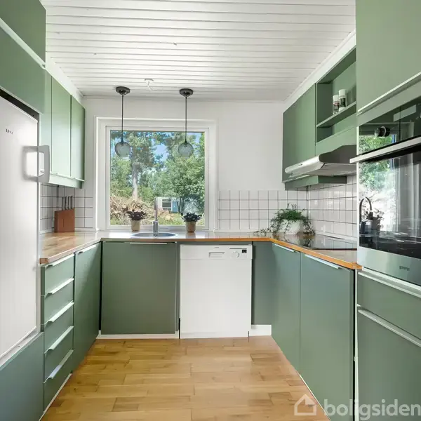 Grønt køkken med moderne apparater, herunder ovn og køleskab. Trægulv og hvidt loft. Stor vindue giver udsigt til grøn have. Planter og lamper dekorerer rummet.