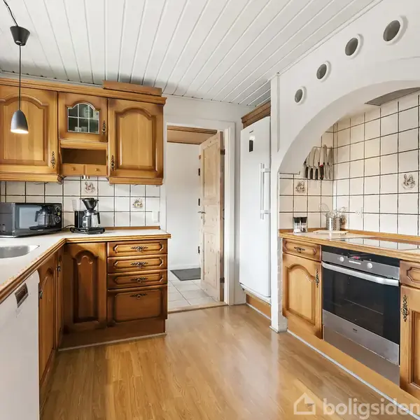 Køkken med træskabe, hvid bordplade, og flisevægge; apparater inkluderer ovn og mikroovn. To pendellamper hænger over bordet. Rummet er lyst med trægulv og hvidt loft.