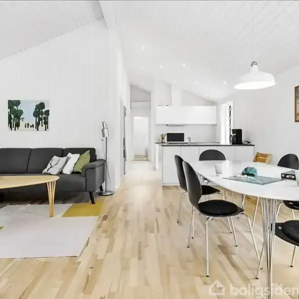 En moderne stue møbleret med en sort sofa og træbord, omkranset af lyse trægulve. Til højre står et spisebord med sorte stole i et åbent køkkenområde, oplyst af hvide lamper.