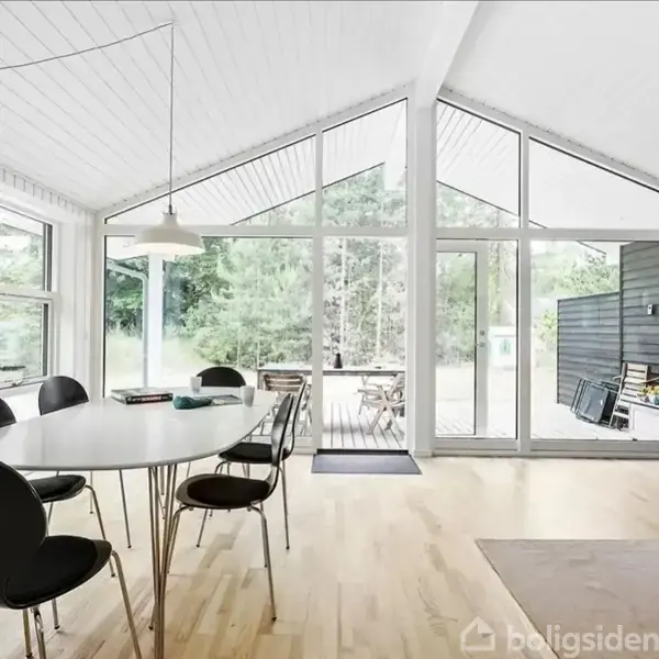 Et moderne spisestueområde med et langt bord og sorte stole. Store vinduer giver udsigt til en terrasse og skov. Lyse trægulve og hvidloftet forstærker rummets lys.