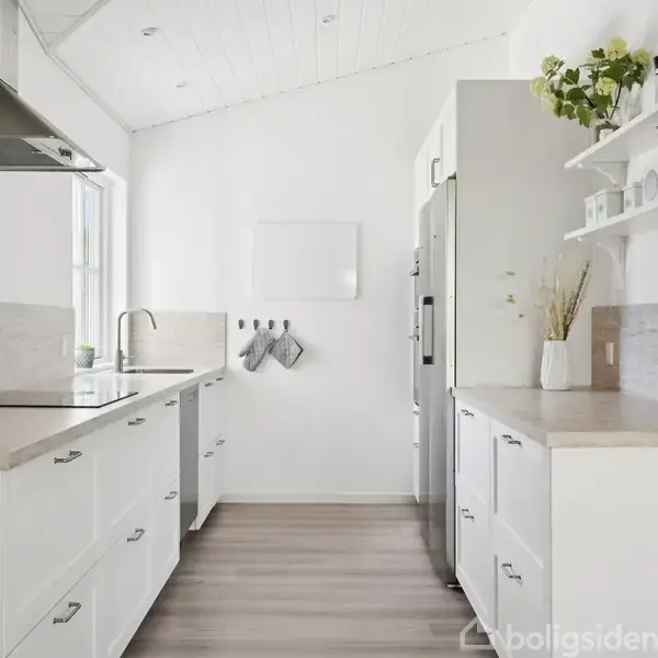 Et moderne, hvidt køkken med indbygget komfurr, vask og stål-emhætte. Der er opbevaringshylder, planter og et køleskab. Loftet er skråt, og gulvet er af lyst træ.