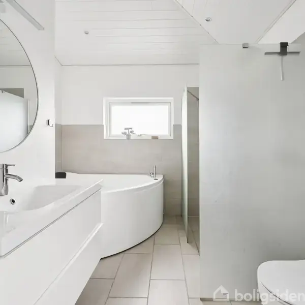 Badeværelse med hvidt badekar og vask i minimalistisk stil. Et stort, rundt spejl hænger over vasken. Der er et vægmonteret toilet og små planter på hylden.