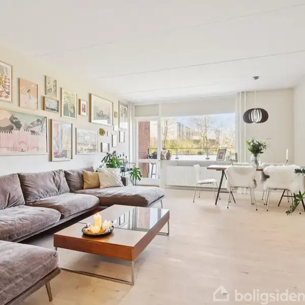 En stor, grå hjørnesofa med puder står i en stue. På væggen hænger mange kunstværker. Foran sofaen er et sofabord, og i baggrunden ses et spisebord nær et stort vindue.