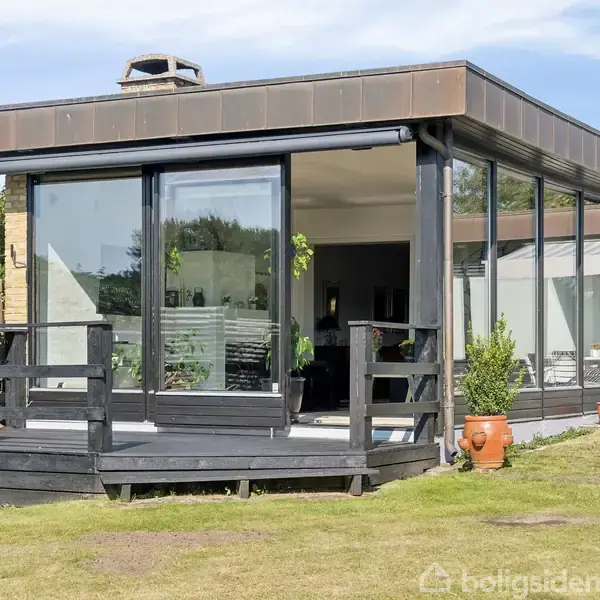 En moderne bolig med store vinduespartier står midt i frodige omgivelser. Huset har en sortmalet træterrasse med potteplanter og grønne buske.