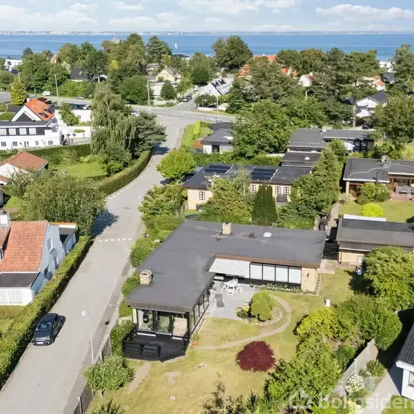 Hus i boligkvarter med have. Beliggende i frodigt område med flere huse og træer omkring. I baggrunden ses et kig mod havet og en klar, blå himmel.