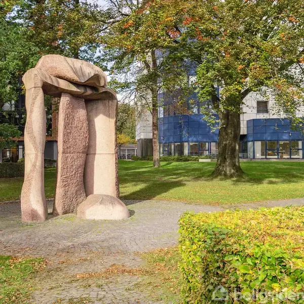 Skulptur i lyserød sten står oprejst, omkranset af græsplæne og træer, med en moderne bygning i baggrunden. Skulpturen har en organisk form og står på en sti af brosten.