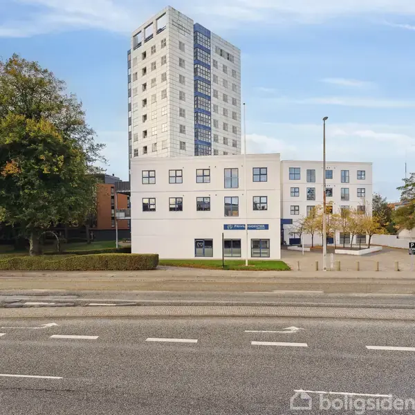 En fleretagers hvid bygning står ved en vej omgivet af træer og en høj bygning i baggrunden. Tekst på skilt: "ROLLING PIN BAKERY."