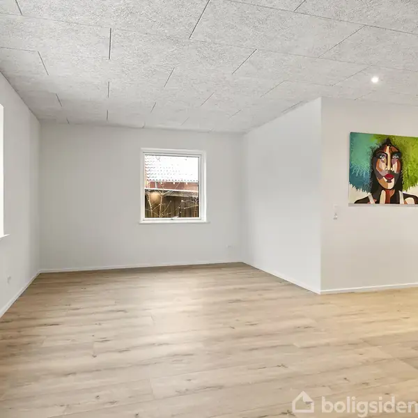 Stue med lyse trægulve og hvidmalede vægge. Et stort vindue og et mindre vindue lukker lys ind. En farverig maleri hænger på væggen.