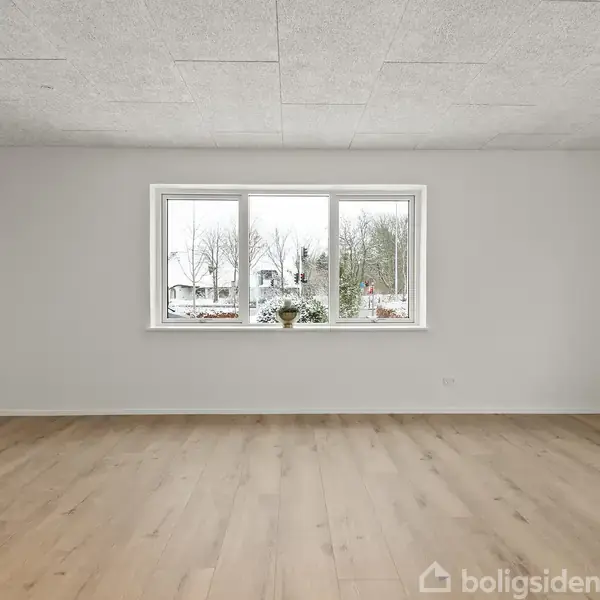 Et rum med lyse trægulve og et stort vindue, der vender ud mod en vinterlandskab med snebeklædte træer. Vinduet har en plante på karmen, og der er hvide vægge.