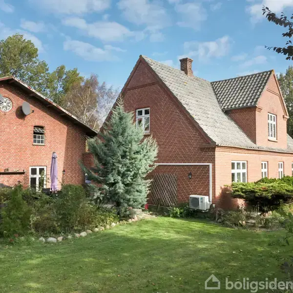 Rødt murstenshus med trekantet tag beliggende i en frodig have. Flere vinduer, to satellitantenner og en lille terrasse er synlige. Græsplæne og buske omgiver bygningen.