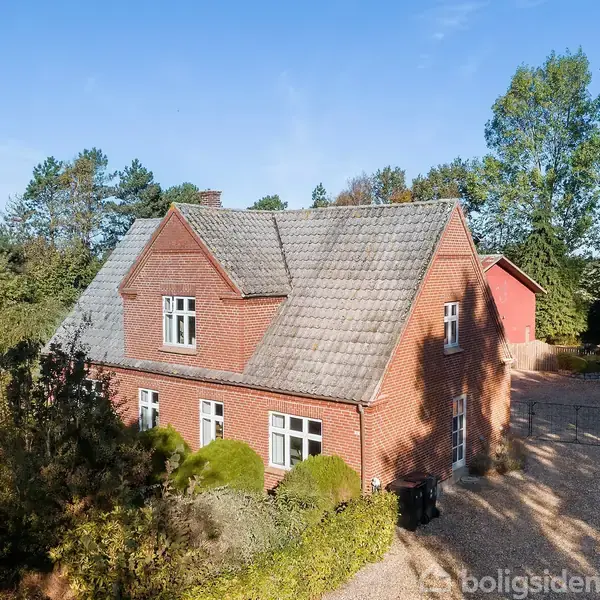 Rødt murstenshus med gråt tag står stille i skovomgivelser omgivet af træer og buske. En anden bygning ses til højre, og gruset indkørsel er synlig.