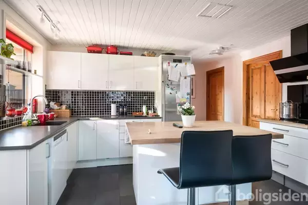 Et moderne køkken med hvide skabe og en sort flisevæg. Et køkkenbord er i centrum, omgivet af to sorte stole. En plante og flere køkkenmaskiner er placeret rundt omkring.