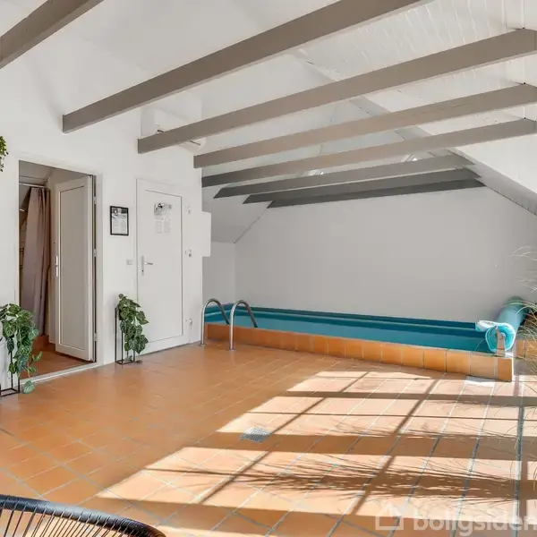 Indendørs pool i en solbelyst rum. Poolen er placeret langs den ene væg, omgivet af planter og møbler. Store vinduer og skråt tag med synlige bjælker.