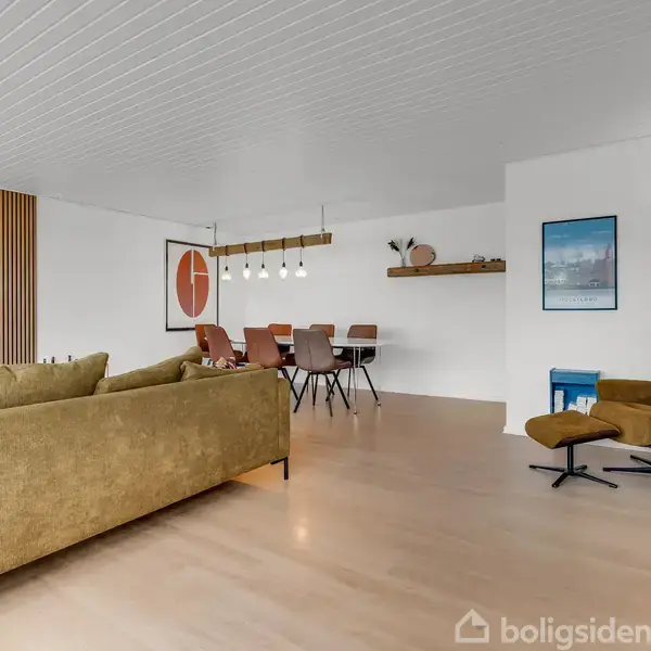 Sofaen står centralt i et moderne stueområde med åbent plas til spisebord. Væggen har dekorativt træpanel, og rummet er oplyst af pendellamper. En lænestol står i hjørnet.