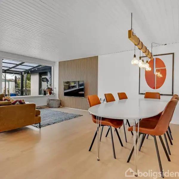Et moderne spisestueområde med et ovalt bord og seks orange stole. Ved siden af er en stue med en brun sofa og et tv på væggen. Store vinduer giver udsigt til en have.