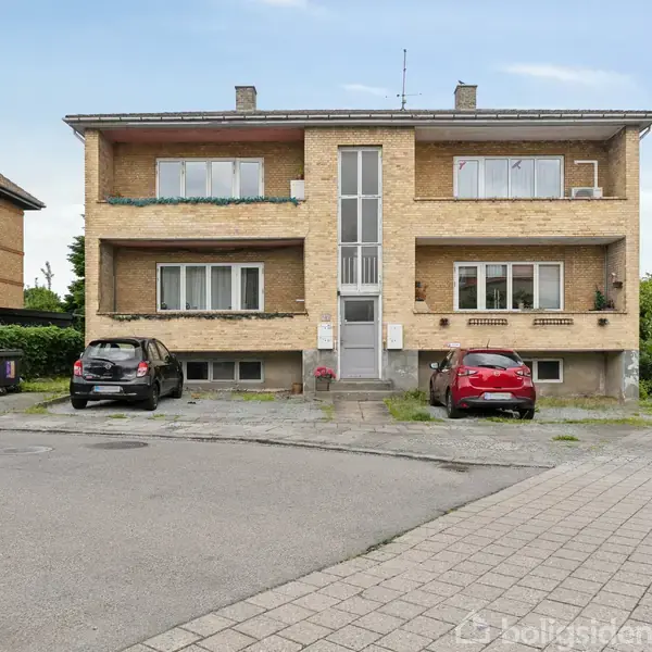 En toetagers murstensbygning med altaner, to biler parkeret foran indgangen, omgivet af grønne buske og en villa til venstre. Asfalteret vej og fortov i forgrunden.