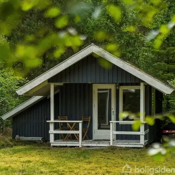 Et lille sort træhus med hvidt tag og veranda står i en frodig skov. To stole og et bord er placeret udenfor.