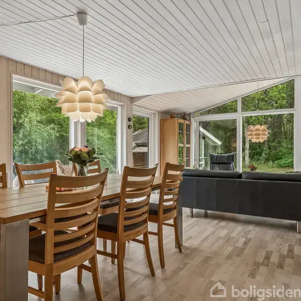 Spisebord i træ med seks stole, står i moderne stue med store vinduer, der viser udsigt til grøn skov. Over bordet hænger en dekorativ lampe. Brændehylde ved siden af sofa.