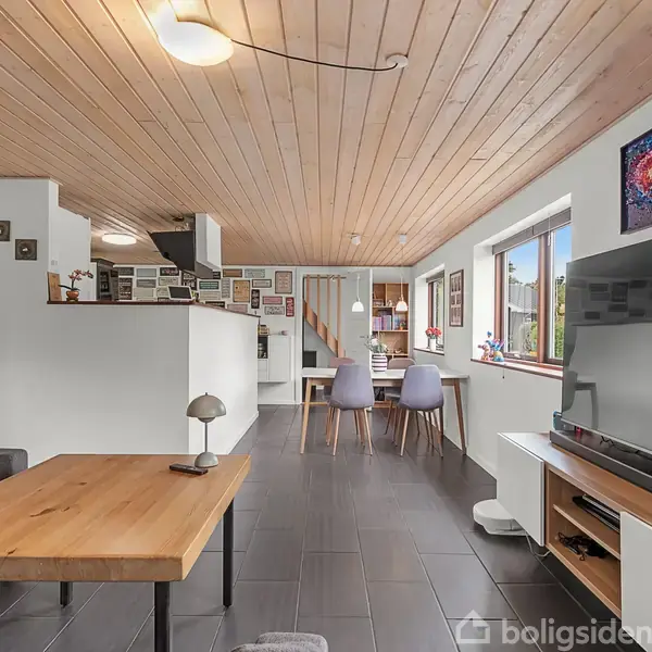 Stue med træloft og sorte flisegulve; sofabord foran sofa; fladskærms-TV på hvid skænk; spisebord med to stole ved vindue; moderne og lys indretning.