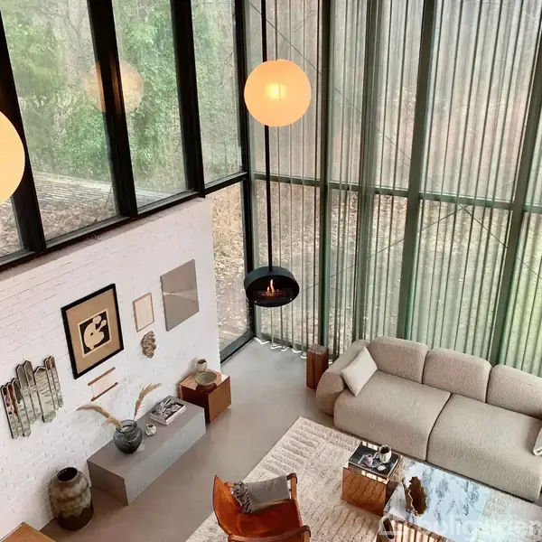 Flydende pejs hænger i glasset stue med beige sofa og kunst på væggen. Store vinduer giver udsigt til naturen udenfor, hvilket skaber en lys og moderne atmosfære.