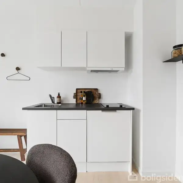 Et lille, hvidt køkken med indbyggede skabe og sort bordplade inkluderer en vask og kogeplade. En plante står på en hylde, og en skammel samt en knagerække ses i baggrunden.