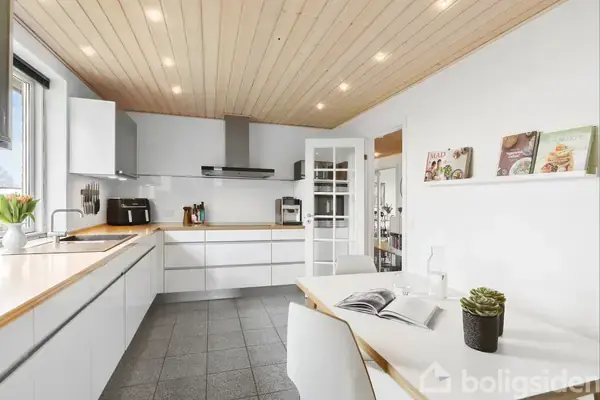 Et lyst køkken har hvide skabe og en spiseplads med åbne kogebøger. En sort emhætte hænger over ovnen, og en lille plante pynter på bordet.