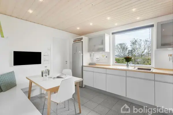 Køkken med spisebord og stole står ved siden af stort vindue med udsigt til træer. Hvide skabe og grå fliser dominerer rummet, med blomster placeret ved vinduet.