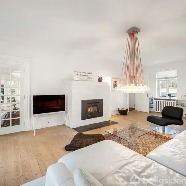 Stor hvid stue med hvid sofa omkring glasbord, åbent ildsted samt moderne lysekrone. Trægulve og store vinduer skaber lys og rummelighed. Dekorationer omfatter billeder og tekst på væggen.