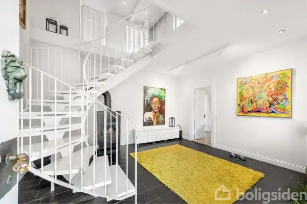 En hvid spiraltrappe står i centrum og fører opad, omgivet af hvide vægge med farverige malerier. En gul løber ligger på sort flisegulv i en lys stue.