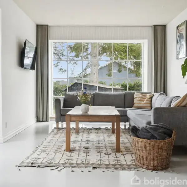 Stue med grå sofaen står ved et stort vindue, omgivet af puder. Et sofabord er centreret på et mønstret tæppe. En kurv og grøn plante er ved siden af.
