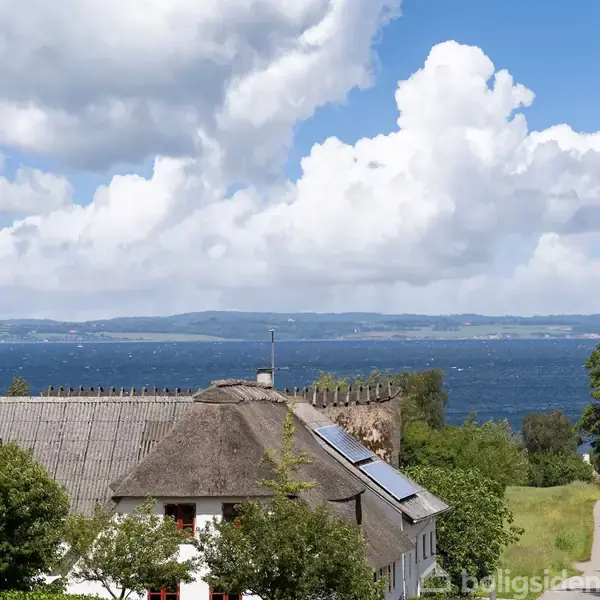 Et bindingsværkshus med stråtag står omgivet af grønne træer, mens det i baggrunden har en udsigt over en blå sø og en himmel med hvide skyer.