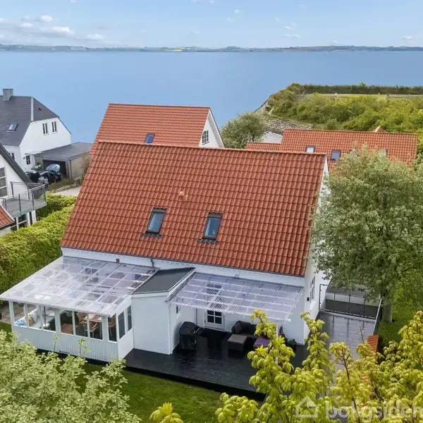 Hus med rødt tegltag og drivhus står i en frodig have ved kysten. Andre huse omgiver det, mens havet strækker sig i baggrunden under en klar himmel.
