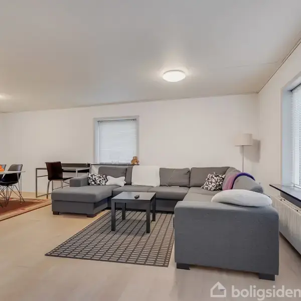 Grå sofa står stille i en moderne stue med lyst trægulv; spisebord med stole er i baggrunden ved et vindue.