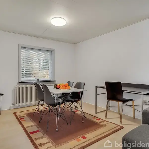 Et bord står dækket med frugt i en lys spisestue. Der er stole omkring bordet, et lille sidebord ved radiatoren og et skrivebord ved vinduet.