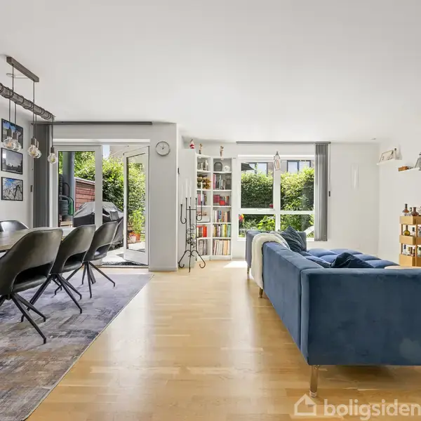 Stue med blå sofa står foran et stort fladskærms-tv. En spisebord med stole er ved siden af. Store vinduer bagved bringer lys ind og giver udsigt til en grøn have.