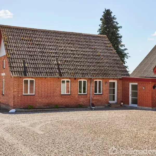 Murstenshus med skråtag står på en grusplads omkranset af landligt landskab, med grønne marker i baggrunden og blå himmel med spredte skyer.