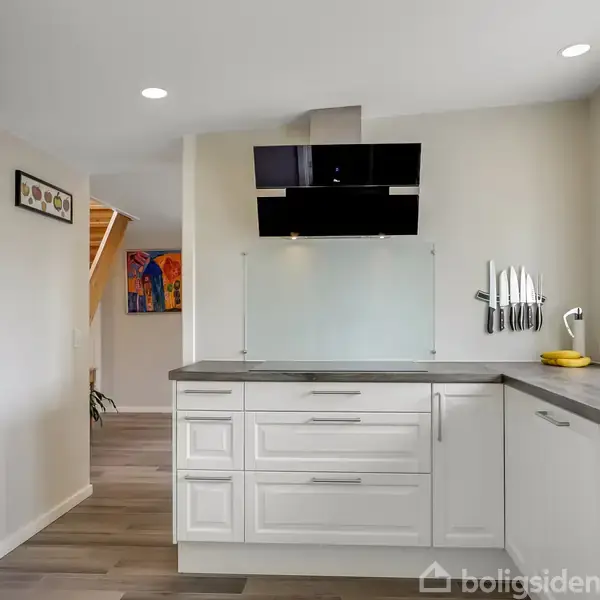 Et moderne køkken med hvide skabe og grå bordplader. På væggen hænger en knivholder, og ved vinduet står en plante. Baggrunden viser en trappe og et farverigt maleri.