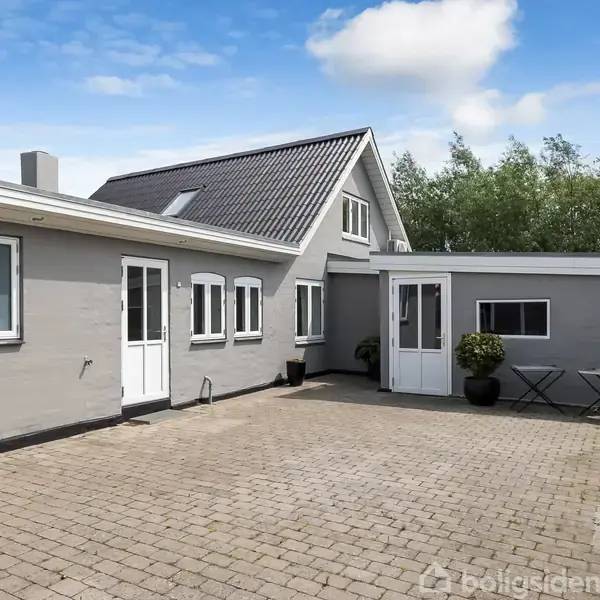 Gråhuset står stille på den flisebelagte gårdsplads, omgivet af grønne træer. To stole og en plante står ved siden af døren. Få skyer ses på den blå himmel.